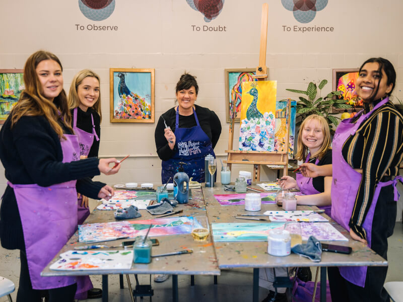 A group of five women in purple aprons smile beside lots of painting materials with a bird painting on an easel
